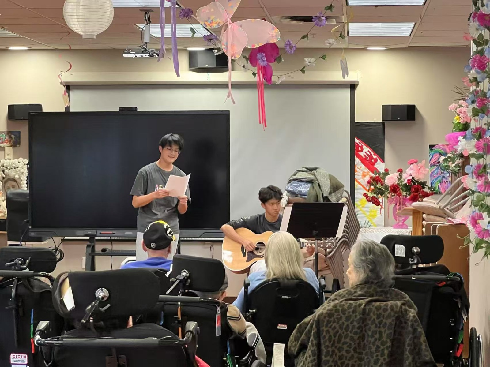 A student volunteer introducing a song while another accompanies on acoustic guitar, residents watching from the front rows