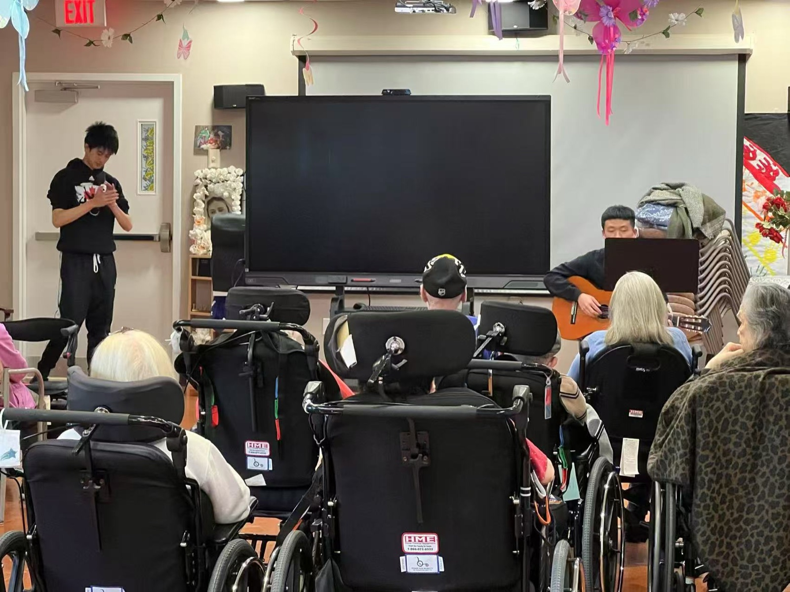 Wide view of a live performance — students on guitar and vocals, residents seated in wheelchairs and chairs