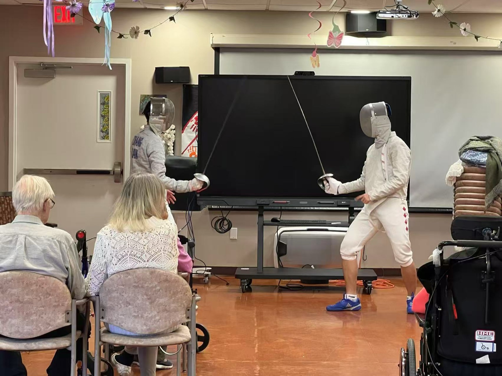 Two student fencers mid-bout, swords crossed, audience seated to the side